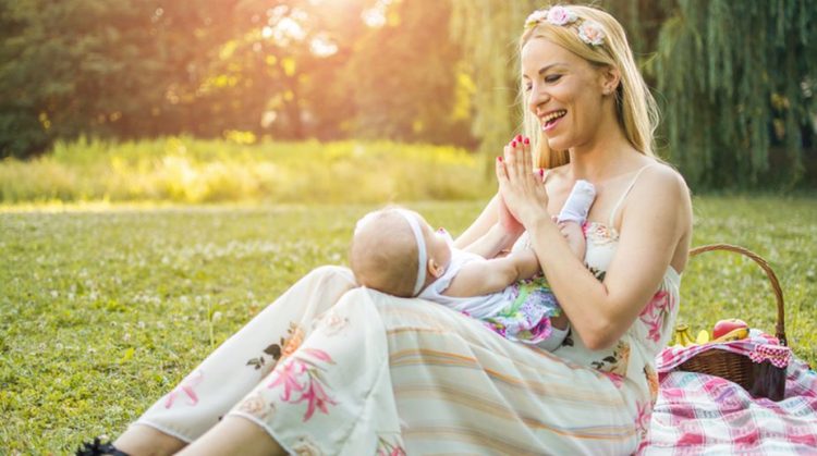 Mother holding baby on lap and clapping hands to make her happy. They are enjoying together beautiful summer day in the nature.